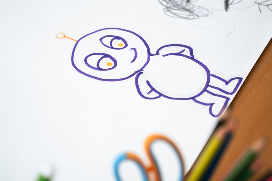 A cartoon character drawn in purple marker on paper, with coloured pencils and scissors beside it